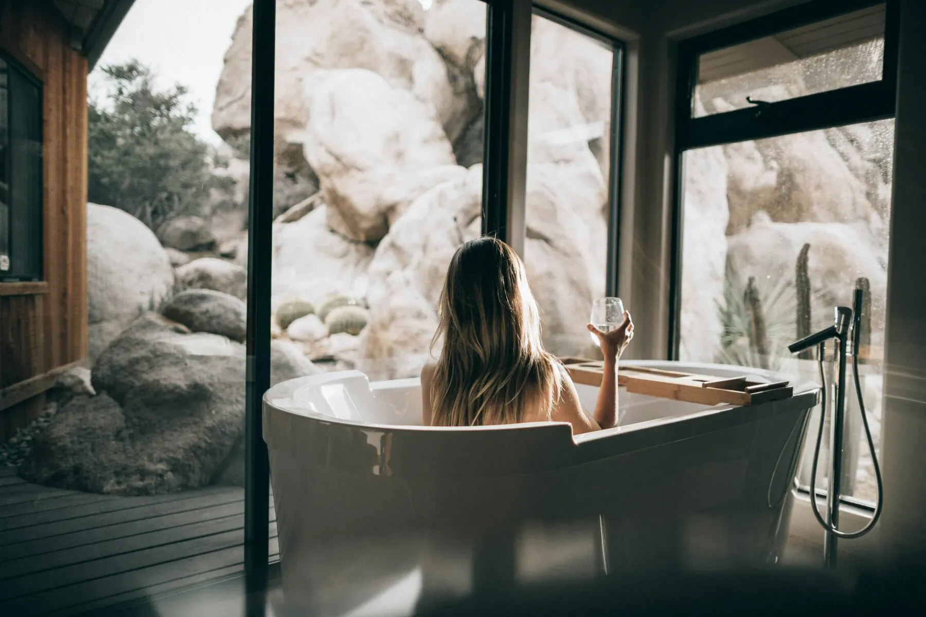 Woman with long blonde hair relaxes nude in white bathtub, holding wine glass, scenic rocky mountain view through window