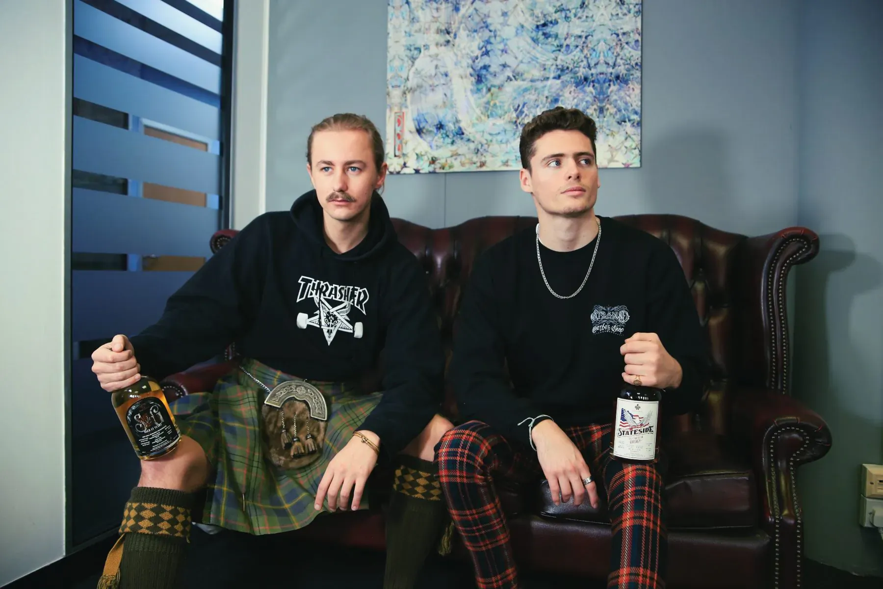 Two Made in Chelsea stars in black hoodies and kilts hold beers on leather sofa, Anti-Burns Night event, abstract art backdrop.
