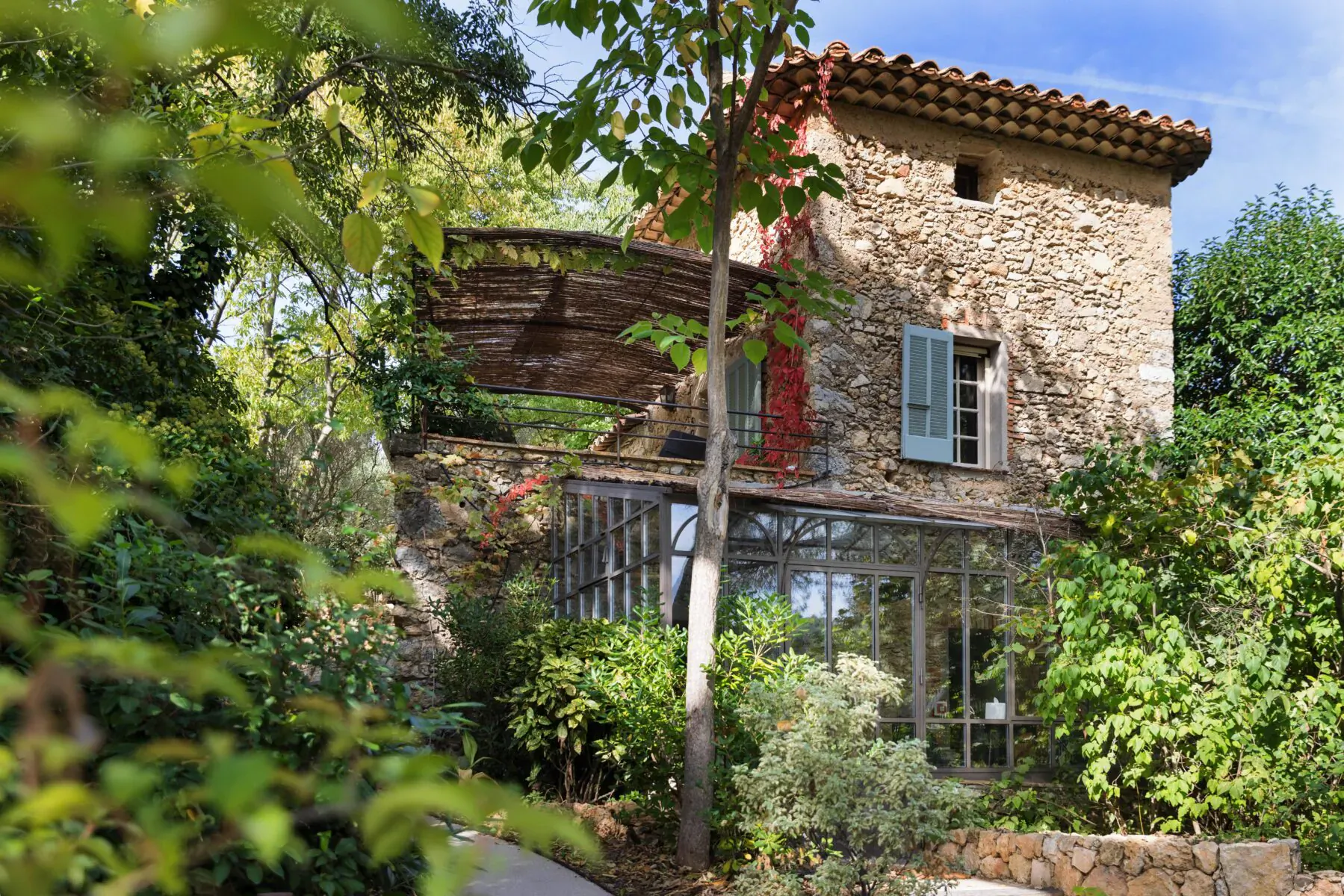 Stone Lou Calen boutique hotel in Provence, with red shutters, greenhouse, surrounded by lush green trees under blue sky