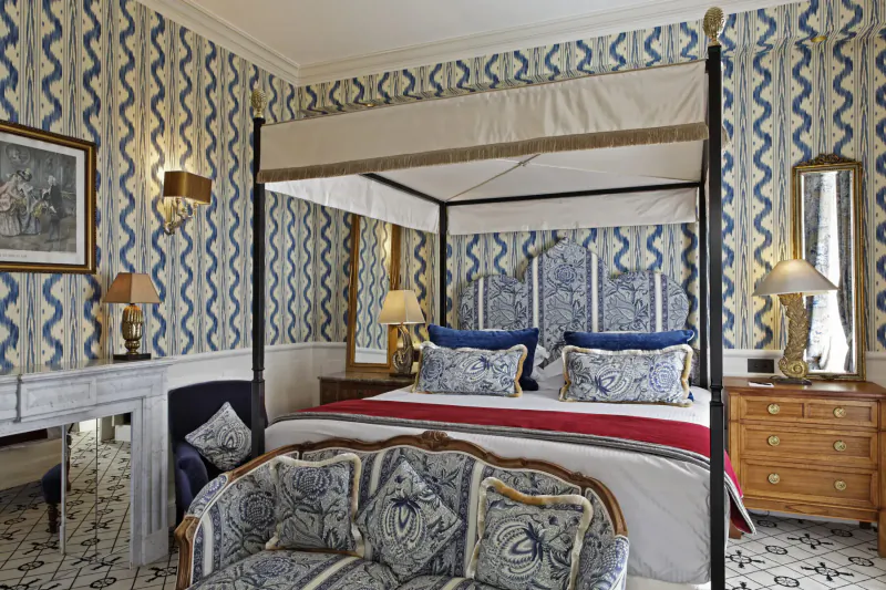 Elegant bedroom in Relais Christine Paris with blue patterned walls, canopy bed, pillows, lamps, and fireplace.
