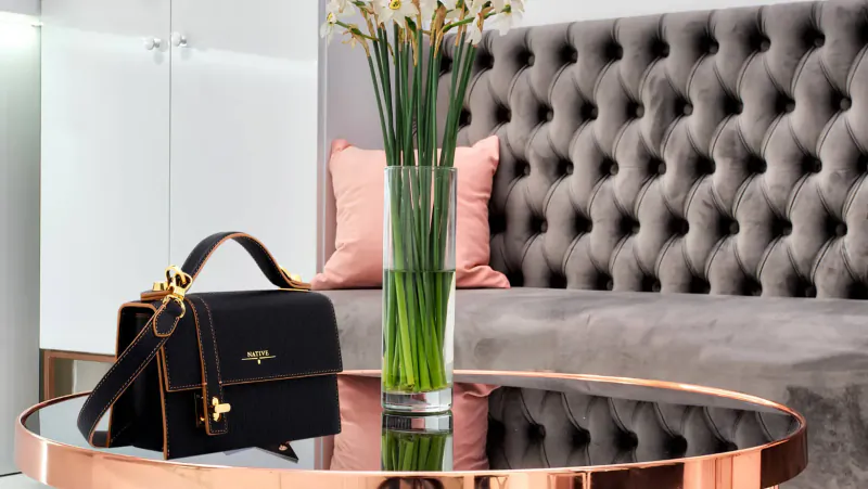 Black designer handbag with keychain on copper-edged glass table beside yellow calla lilies in pink vase on gray tufted sofa