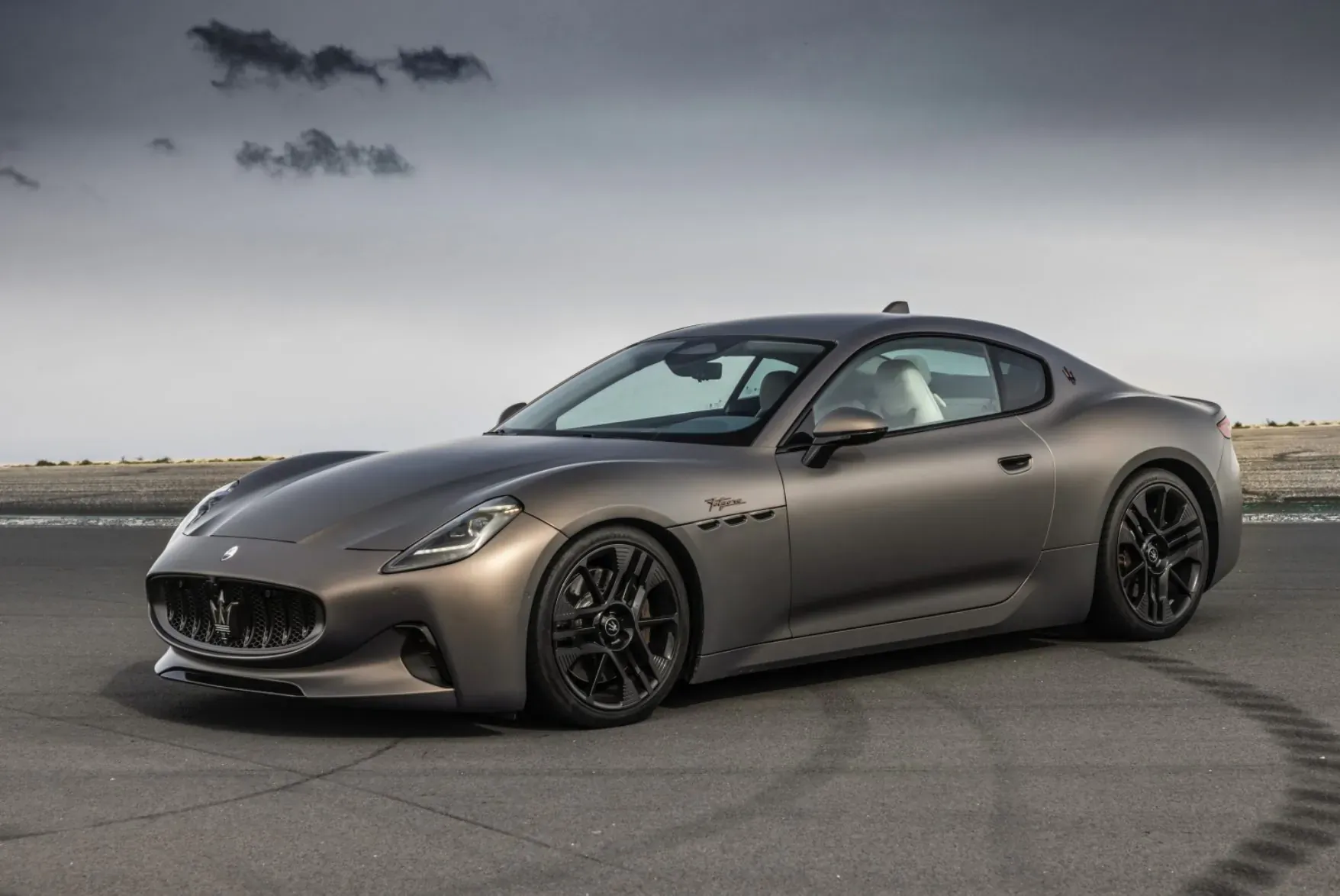 Metallic gray Maserati GranTurismo Folgore coupe on racetrack, tire marks, cloudy sky.