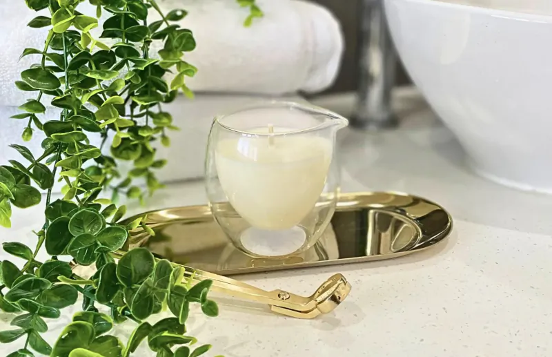 Glass jar with creamy Amber Hue candle on gold tray with tweezers, green foliage, white towels, and sink nearby
