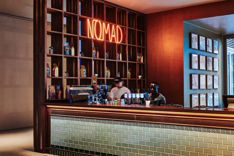 Nomad bar at 25hours Hotel Dubai with neon sign, wooden shelves of bottles, bartenders at counter, warm lighting.
