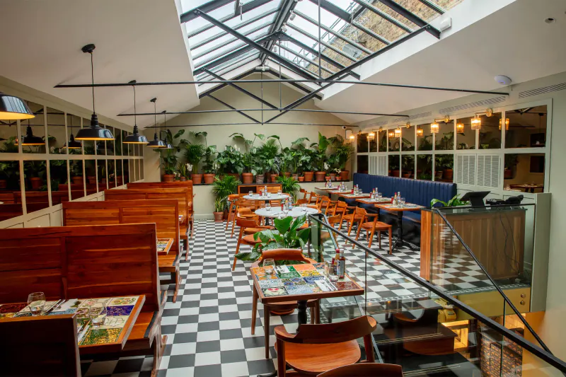 Spacious Lobster Pot Booth at The Mayfair Chippy: greenhouse-style dining room with skylights, plants, wooden booths, checkered floor, set tables.