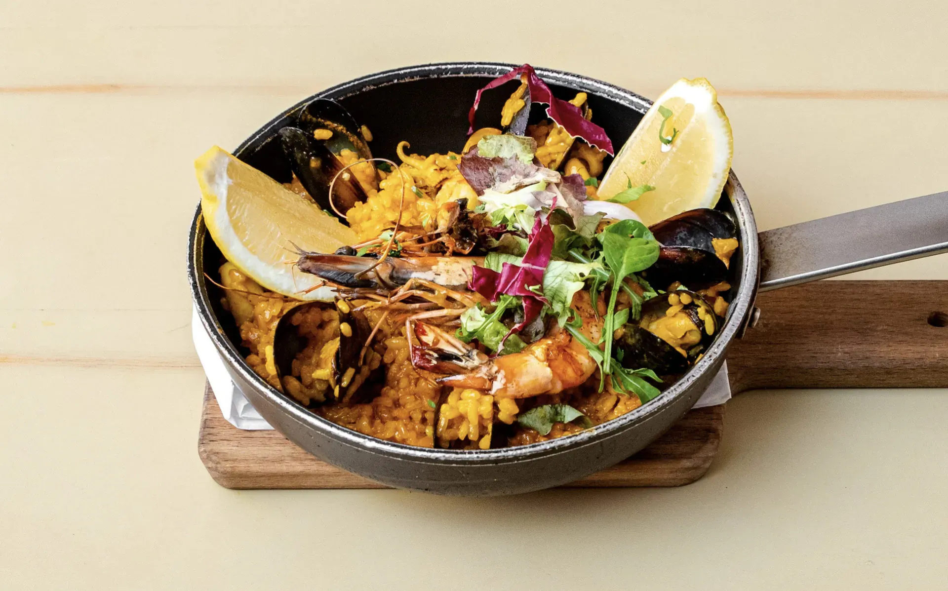 Paella with mussels and prawns in black pan on wooden board, garnished with lemon and herbs