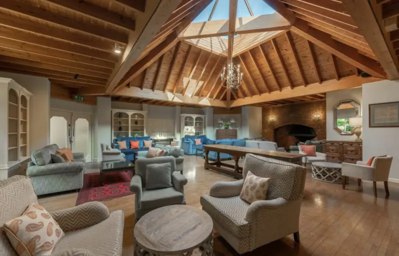 Spacious luxurious hotel lounge with wooden beamed ceiling, skylight, chandeliers, plush sofas, tables, and fireplace.