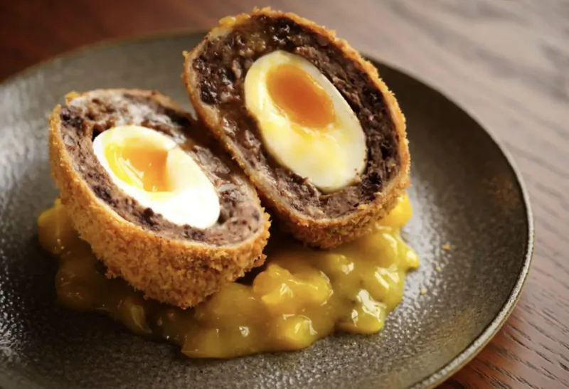 Two halved Scotch eggs with runny yolks on yellow mustard sauce on a dark plate