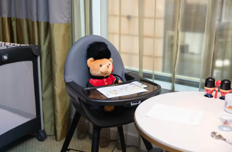 Paddington Bear in red uniform sits in black high chair at Guardsman hotel room table with toy guards and window view