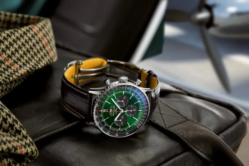 Breitling Navitimer green dial chronograph watch on leather strap, on brown leather bag with tweed jacket and airplane propeller backdrop