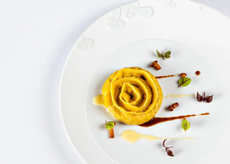 Swirled yellow pasta on white plate garnished with microgreens, drizzled sauces, Michelin-style.