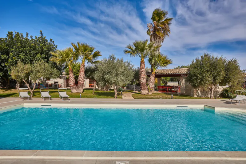 Luxury villa's turquoise infinity pool surrounded by palm and olive trees under blue sky, Sicily.