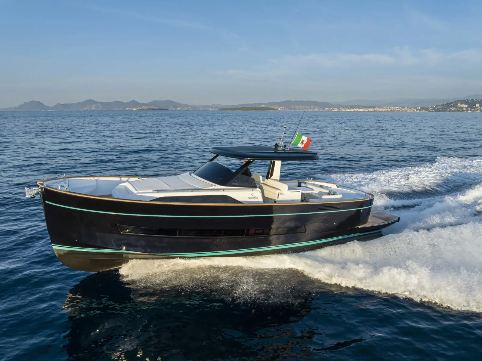 Black luxury motor yacht with Italian flag speeding on sea, distant mountains and city skyline visible.