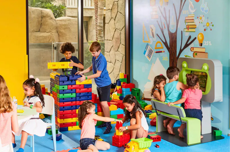 Diverse children building with colorful LEGO bricks and playing at tables in a bright hotel kids' playroom with educational wall mural.