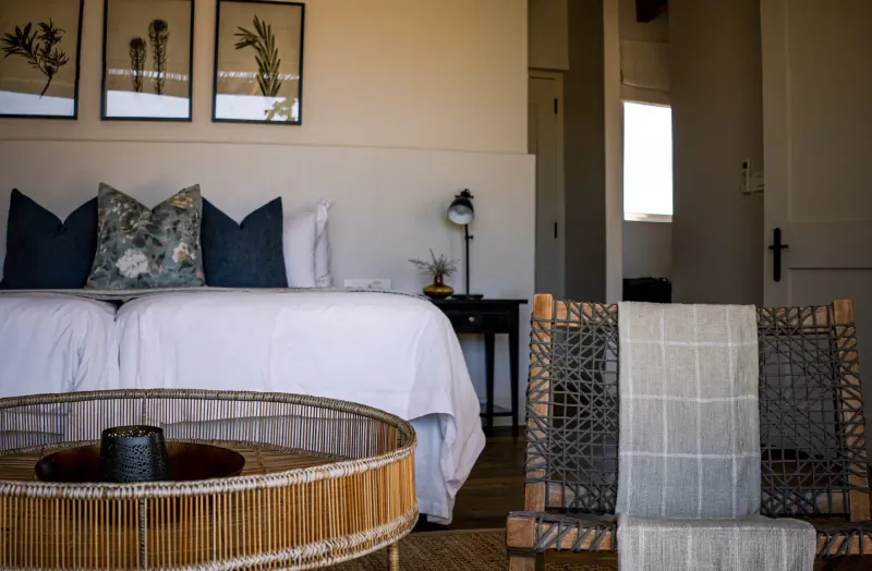 Cozy bedroom retreat with white bed, patterned pillows, rattan table, chair with throw, botanical art, and lamp in modern cabin.
