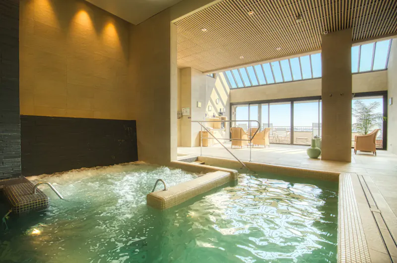 Indoor hot tub with bubbling turquoise water in modern spa lounge, large windows, chairs, and sea view.