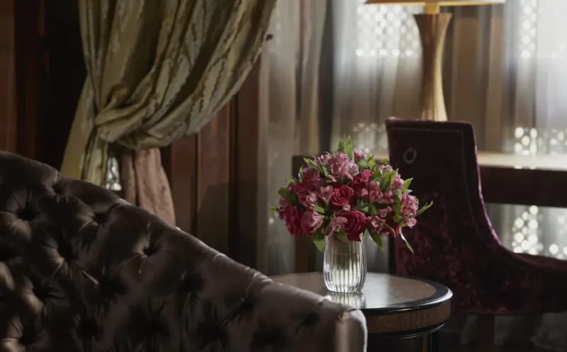 Luxurious hotel room with pink roses in glass vase on round table, tufted armchair, velvet chair at desk, green drapes, lamp, window.