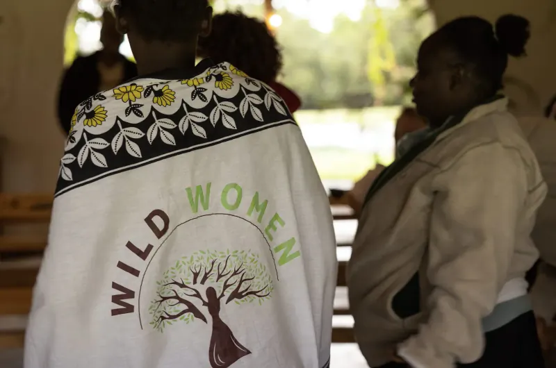 Group of Black women wearing 'WILD WOMEN' shawl with tree logo, standing outdoors under arches near park.