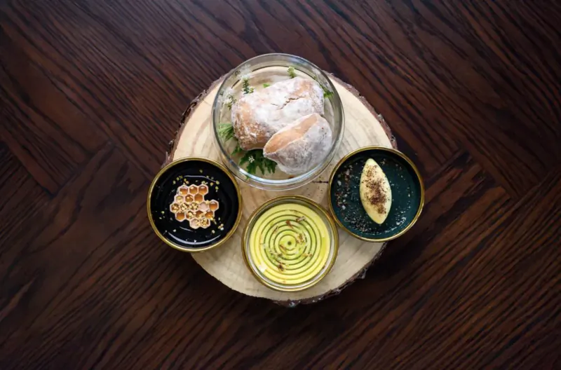 Foraged dish on wooden board: poached fish in glass bowl with greens, black honey pot, yellow swirl sauce, and pastry in dishes