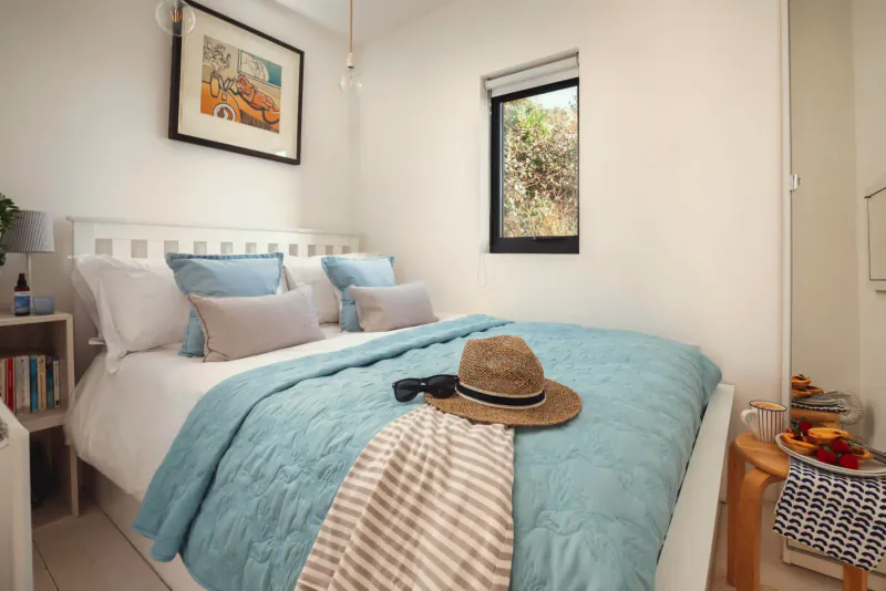 Cozy boutique bedroom with turquoise bedding, straw hat, sunglasses, books, fruit bowl, and beach art.