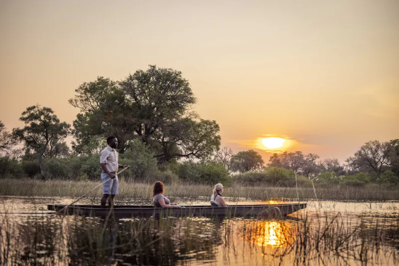 Man fishing from canoe with two women at sunset in African safari wetlands amid acacia trees