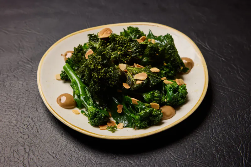 Plate of steamed broccoli with almonds and sauce dots on dark background