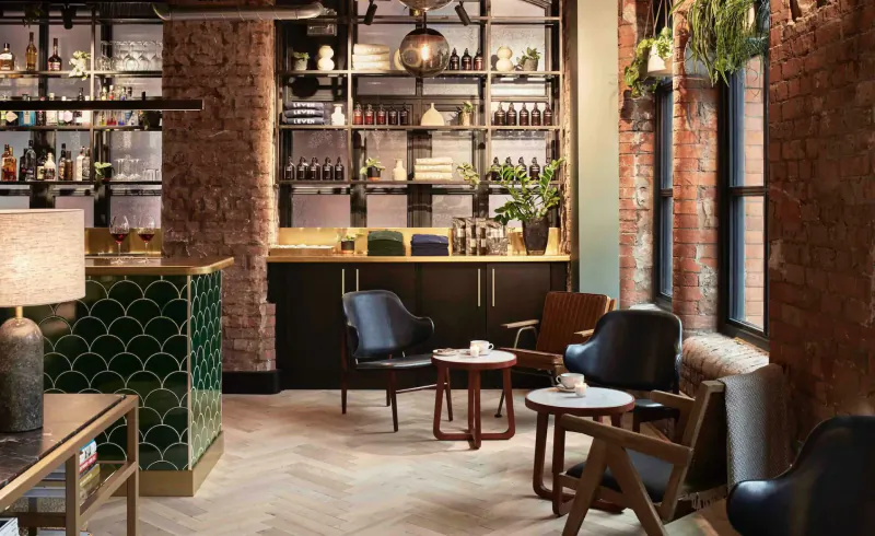 Stylish Leven Manchester lounge with exposed brick walls, green bar, wooden chairs, plants, and pendant lights.