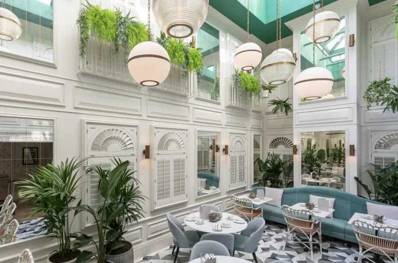 Elegant atrium interior of 100 Queen's Gate Hotel with white walls, green plants, globe lamps, teal ceiling, and cafe seating.
