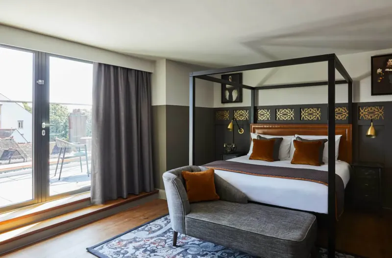 Elegant Hotel Indigo Chester bedroom with black four-poster bed, orange pillows, gray ottoman, and balcony view.