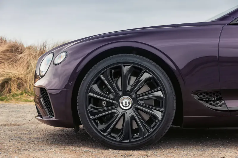 Purple Bentley GT Mulliner Blackline with black wheels, front side view on coastal path amid tall grass.