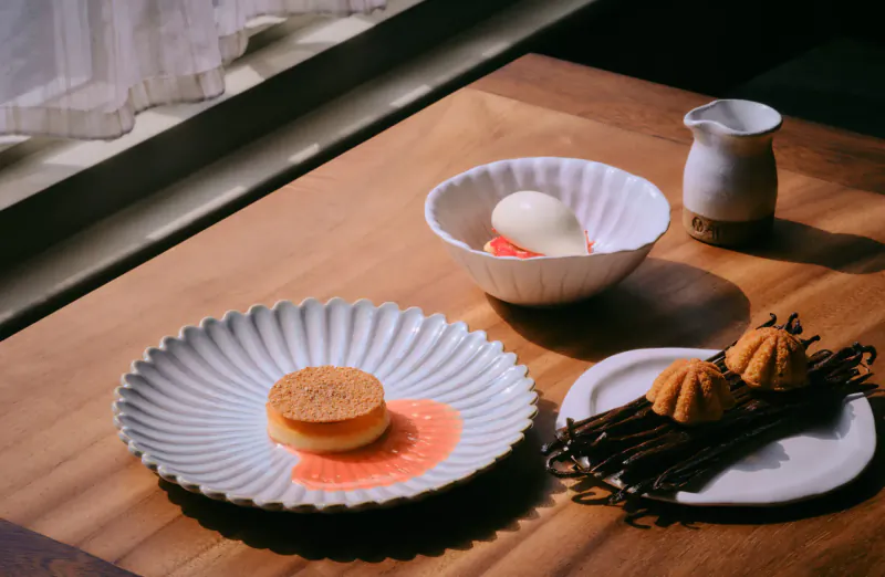 AngloThai Restaurant: flan with orange sauce on scalloped plate, poached egg in bowl, milk jug, vanilla pods on table by window.