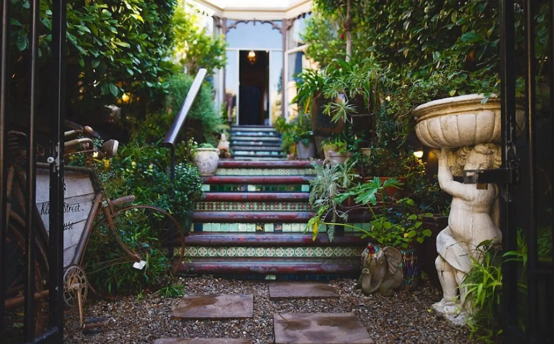 Lush garden courtyard with ornate tiled steps leading to open door, cherub statue, plants, and vintage elements on Kloof Street, Cape Town.