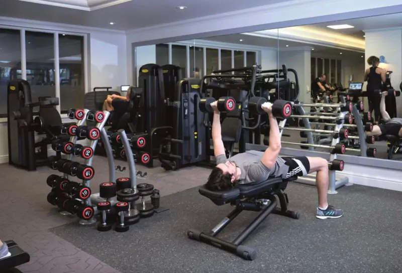 Man bench pressing dumbbells on incline bench in modern hotel gym with mirrors and equipment.