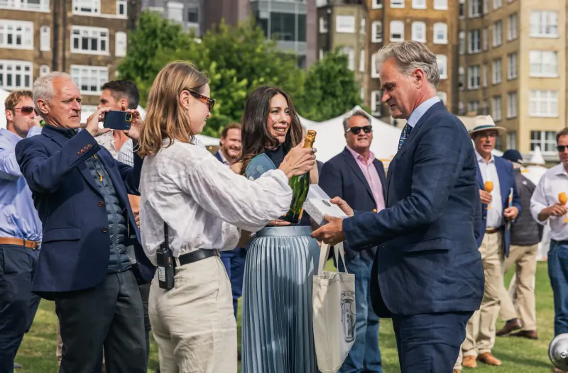 Woman in blue dress toasting with champagne bottle to man in suit at London Concours 2024 event amid crowd and classic cars.