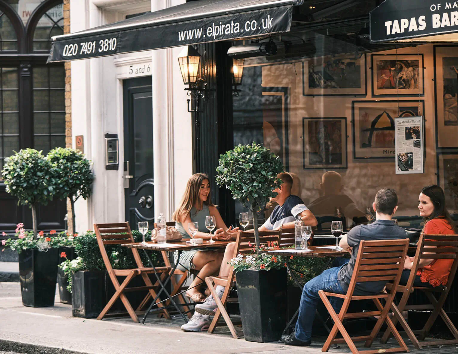 El Pirata Tapas outdoor patio in Mayfair: diverse group at wooden table with wine glasses, www.elpirata.co.uk sign, potted plants.