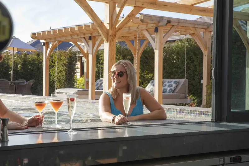 Blonde woman in bikini and sunglasses at poolside bar, holding champagne flute, Manor House Hotel spa pergola view
