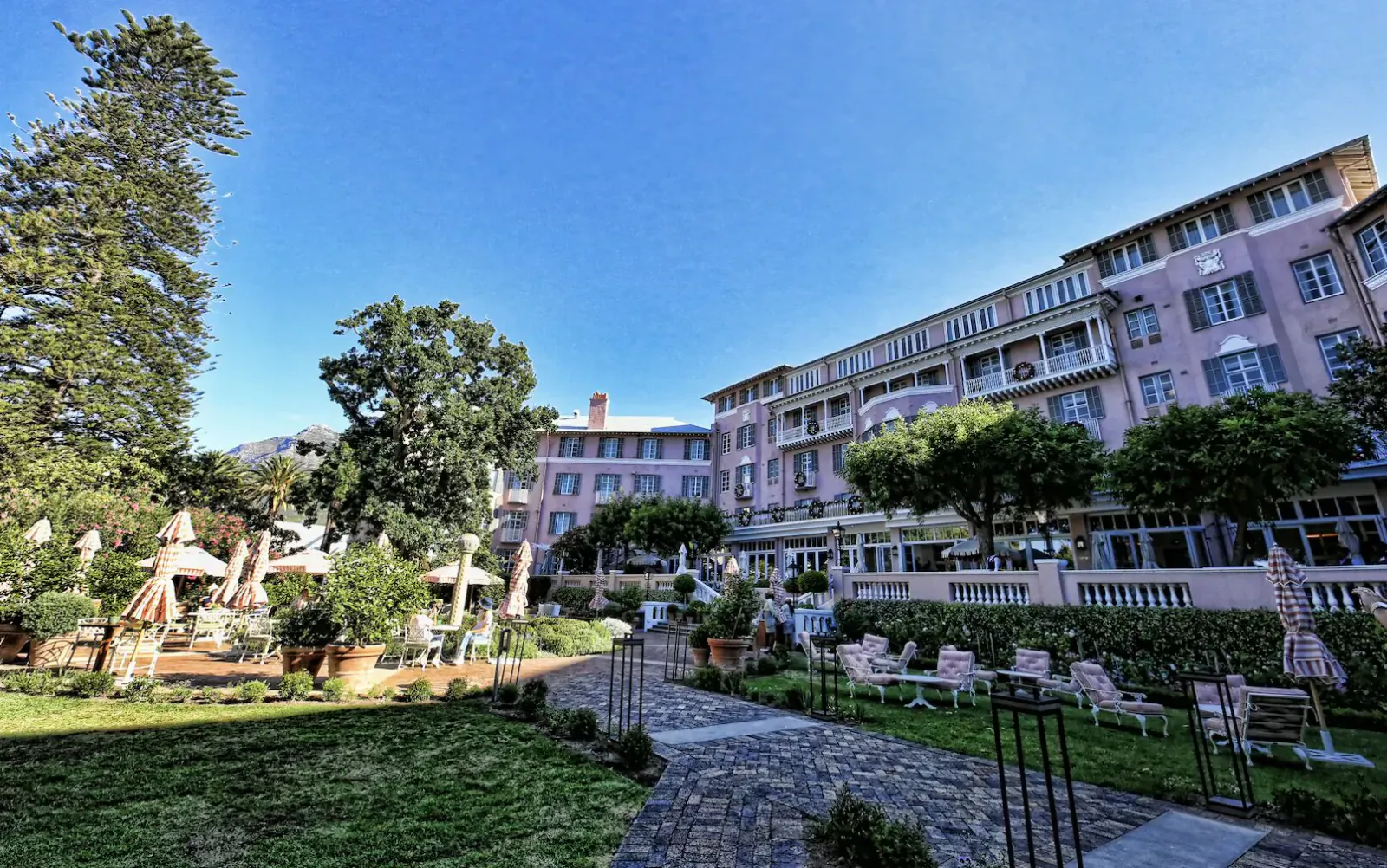 Pink Mount Nelson Hotel in Cape Town with lush gardens, palm trees, lounge chairs, and umbrella tables under blue sky