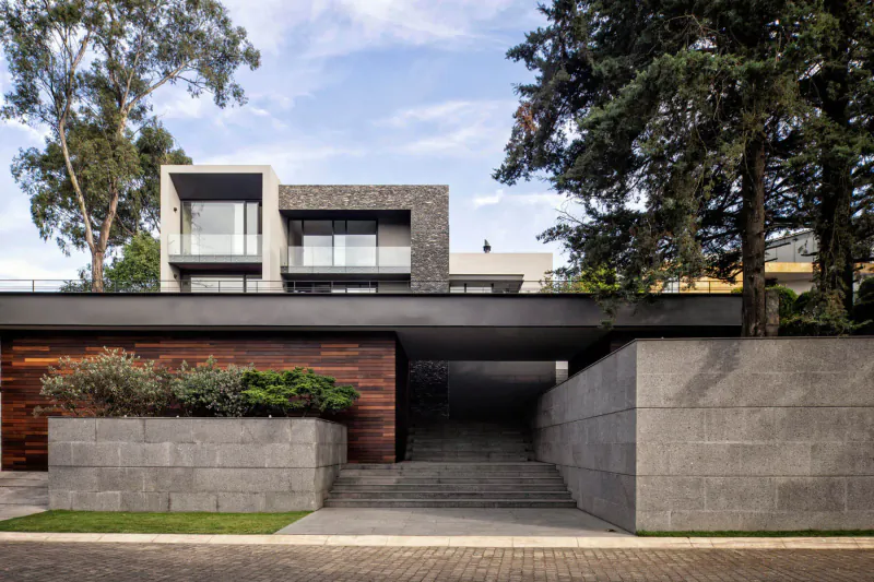 Modern Mexican house with concrete, wood, and glass facade, surrounded by trees and landscaped entrance under blue sky.
