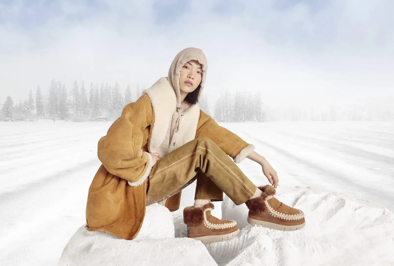 Model in beige shearling hooded coat, pants, and UGG-style boots sits on snowy rocks amid winter forest backdrop