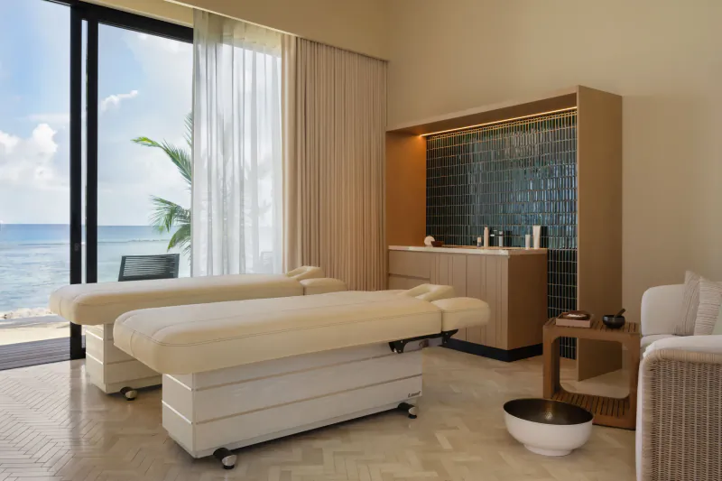 Luxurious spa room at SO/ Maldives with two white massage tables, wooden cabinet, and ocean view through glass doors.