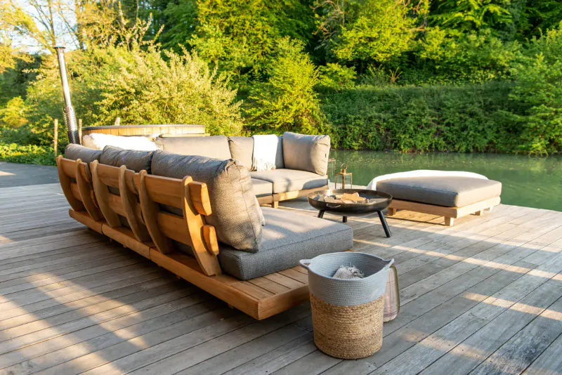 Modern wooden outdoor sofa set with gray cushions, coffee table, ottoman, and basket on deck by lush green garden.