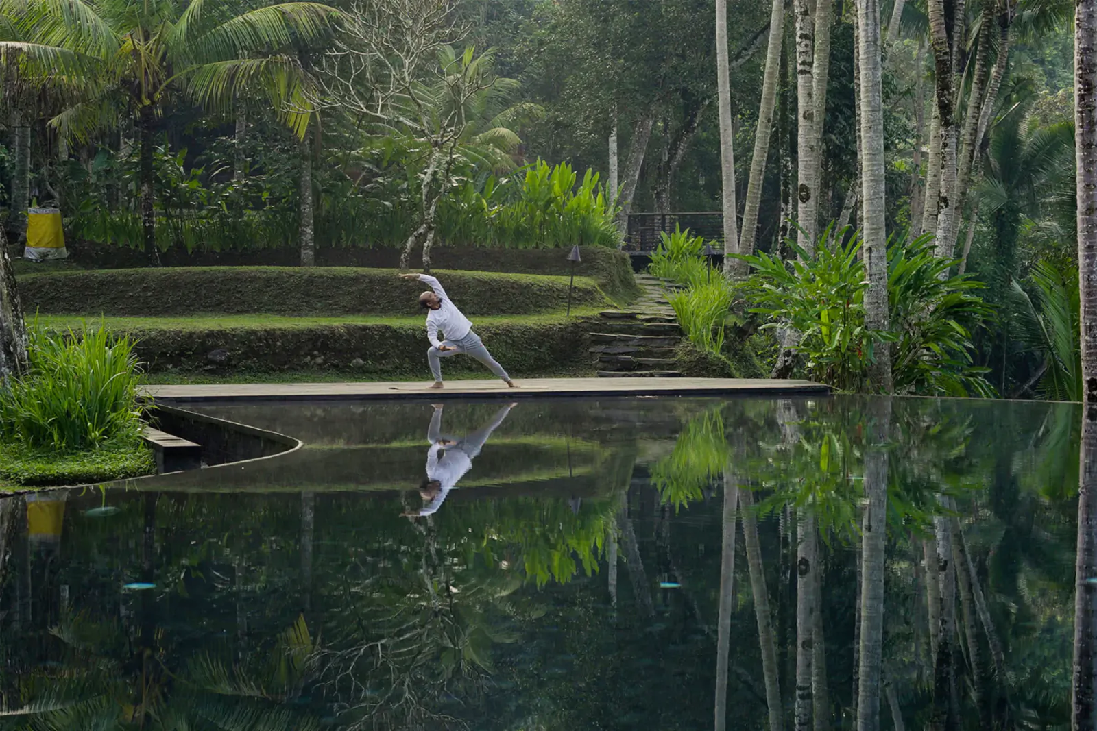 Person in white performs yoga pose on wooden dock over reflective pool in lush tropical forest, Bali.