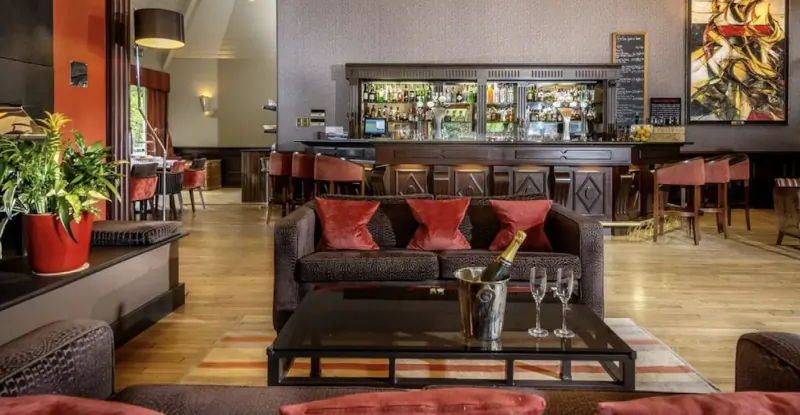 Luxurious hotel lounge with red sofas, wooden bar, champagne bucket with glasses, plants, and abstract art.