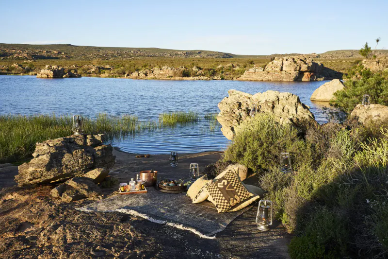 Luxurious picnic setup with lanterns and food on rug amid rocks, reeds, and serene lake at Bushmans Kloof wilderness reserve