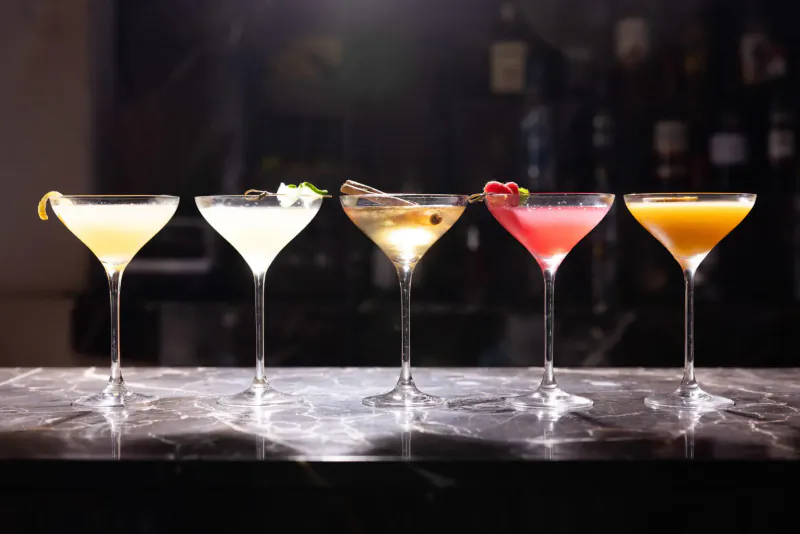 Five elegant cocktails in martini glasses on a marble bar: lemon, white flower, gold snake fruit, raspberry pink, orange, with blurred bottles behind.