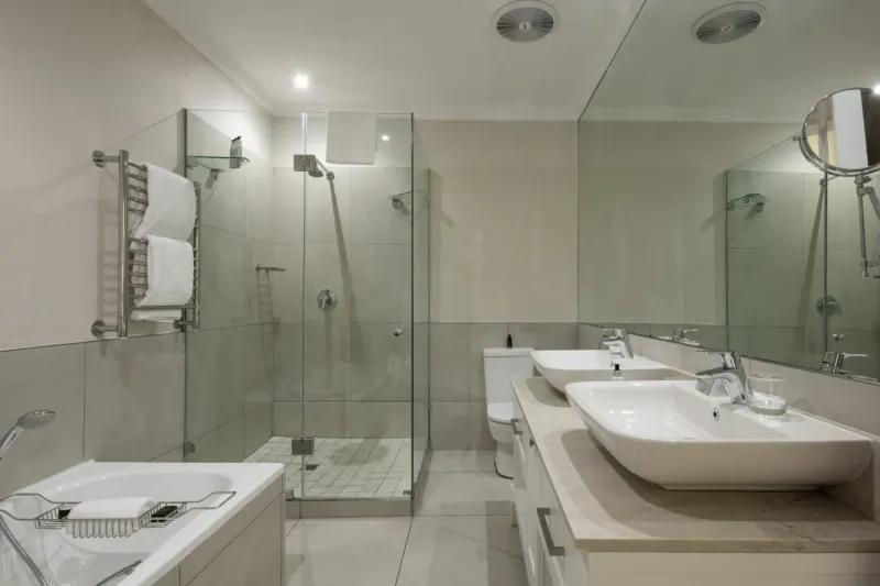 Modern hotel bathroom with glass shower, double white sinks, towel warmer, and mirrored walls.