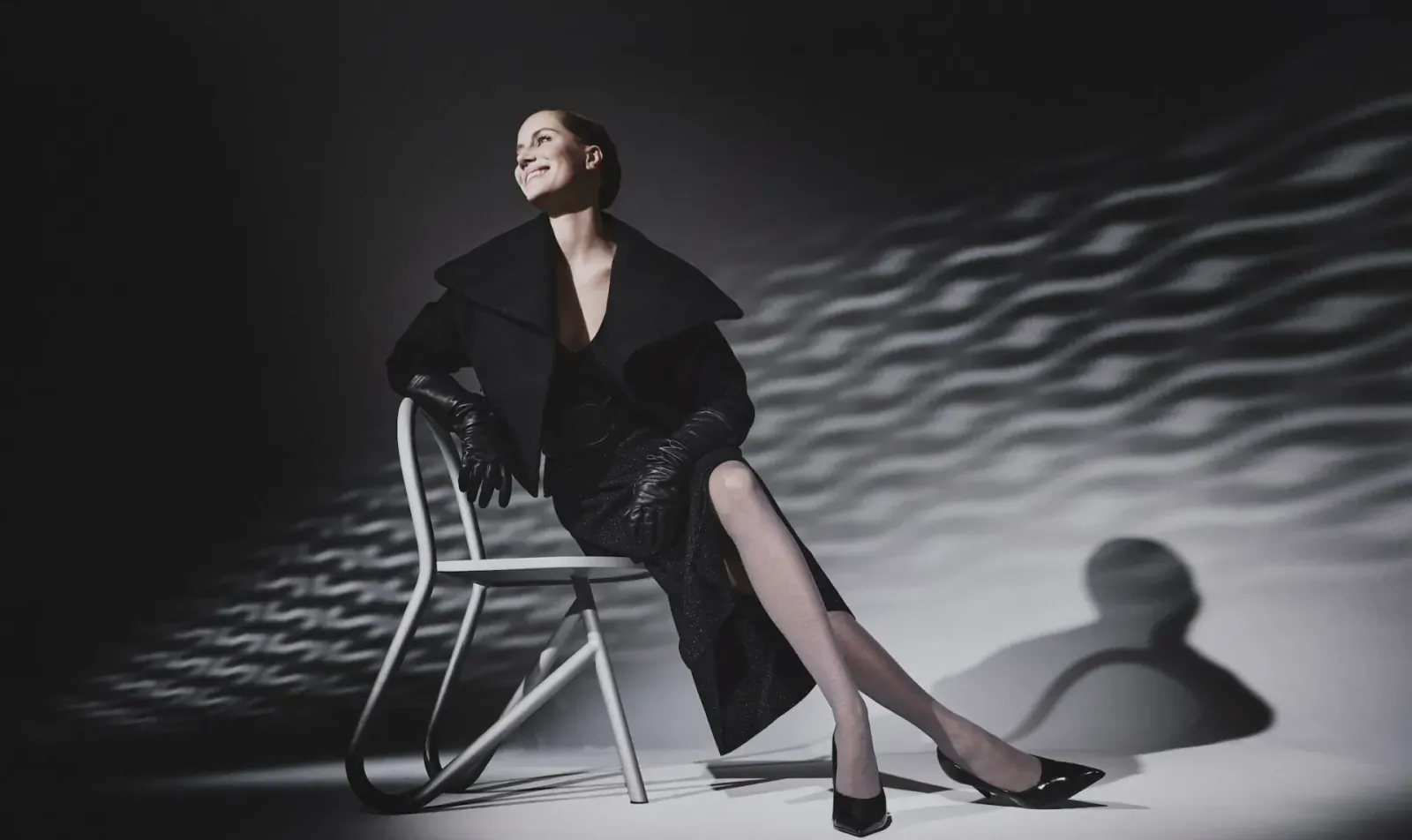 Elegant woman in black fur-trimmed dress and gloves, legs crossed on metal chair, dramatic light patterns and shadows, for FALKE 130 years.