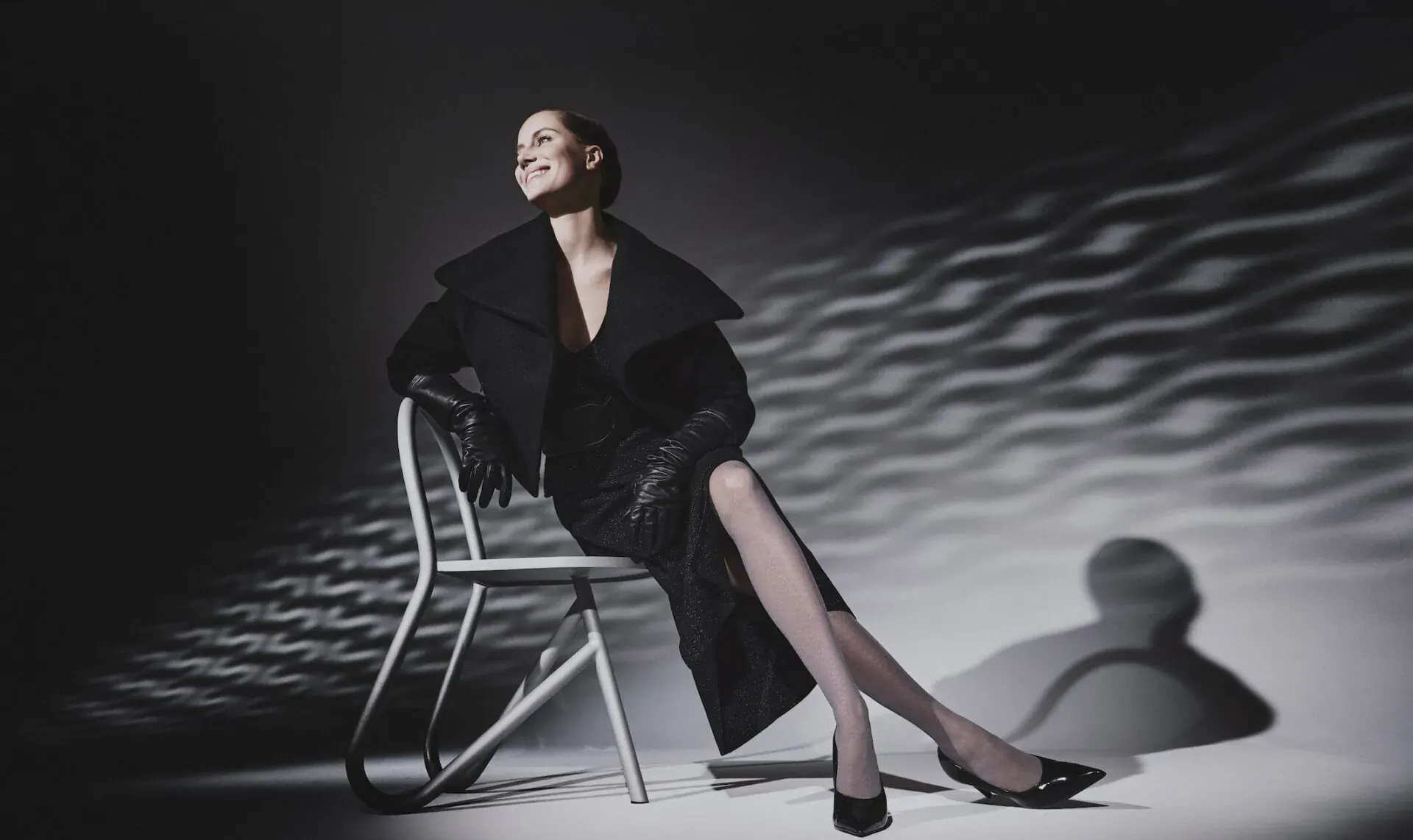 Elegant woman in black fur-trimmed dress and gloves, legs crossed on metal chair, dramatic light patterns and shadows, for FALKE 130 years.