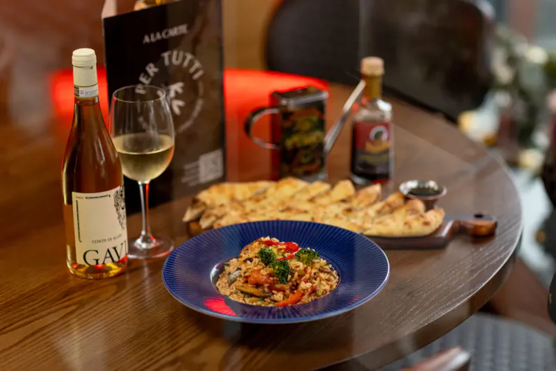 Per Tutti table setting with Gal wine bottles, white wine glass, blue bowl of pasta with peppers, bread, and dips