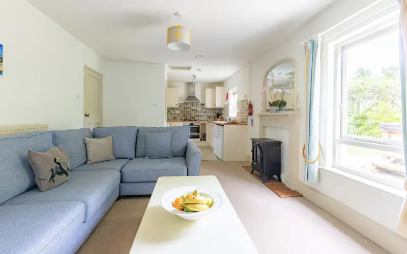 Bright living room with blue L-shaped sofa, white coffee table with bananas, open kitchen, and window garden view at Broomhill Manor.